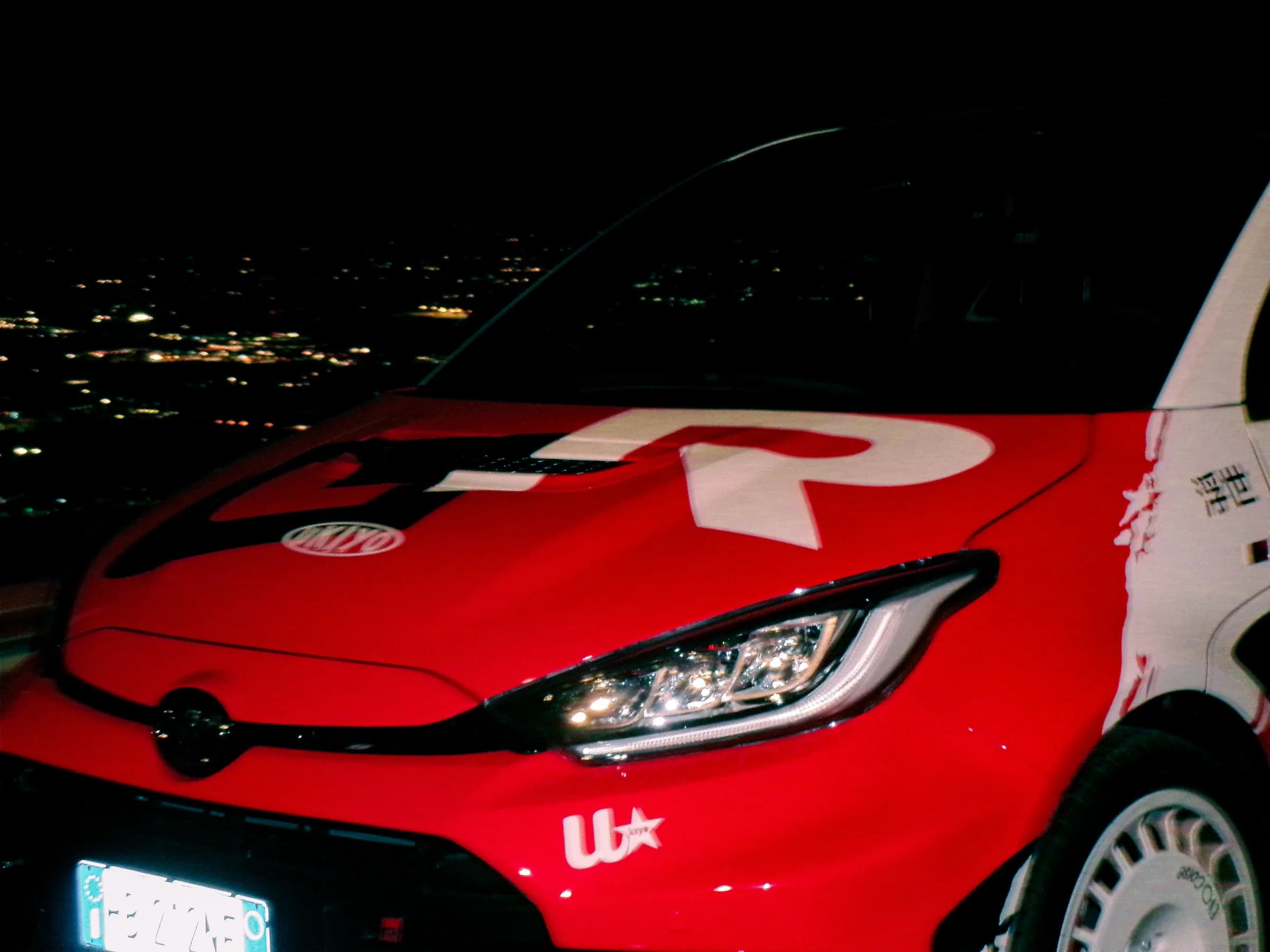 Una Toyota GR Yaris con livrea rossa e bianca fotografata di notte con le luci della città sullo sfondo.