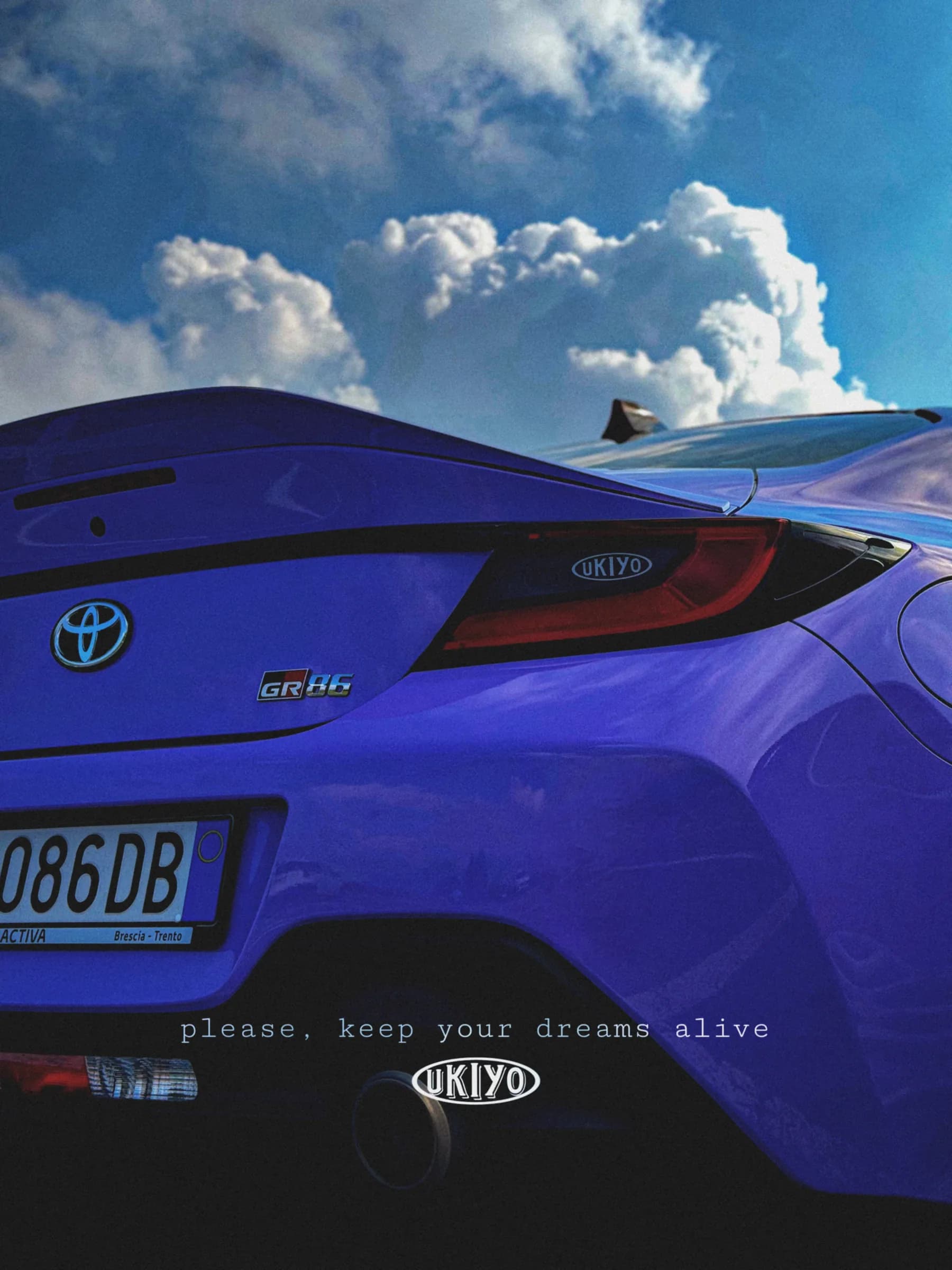 Una Toyota GR86 blu fotografata dall'alto contro un cielo di nuvole, con la scritta “please, keep your dreams alive”.
